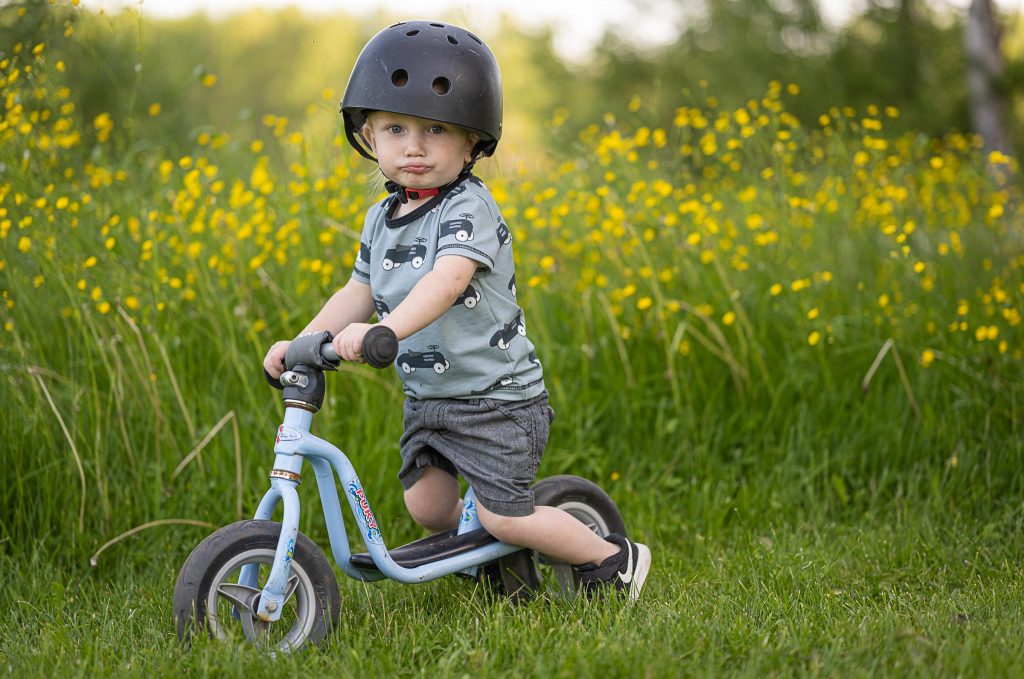 Pieni poika istuu potkupyörän päällä. Hänellä on kypärä päässä ja taustalla on keltainen kukkameri.